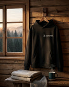 Hoodie Höhenlage in Black mit Todtnauberg-Brustdruck, am Holzbügel in einer Berghütte vor dem Fenster mit Blick auf den Schwarzwald