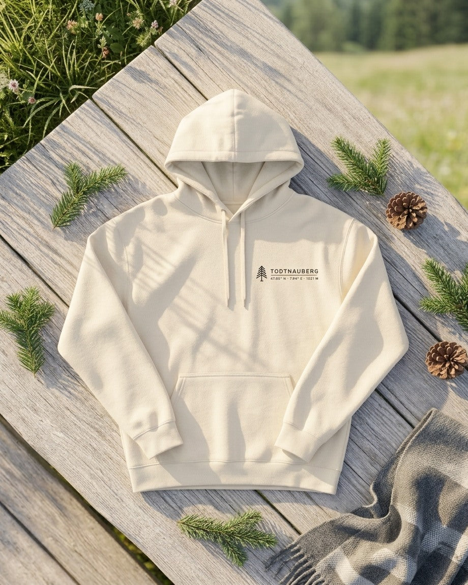 Hoodie Höhenlage in Natural Raw mit Todtnauberg-Brustdruck, ausgebreitet auf einer Holzbank mit Tannenzweigen und Zapfen